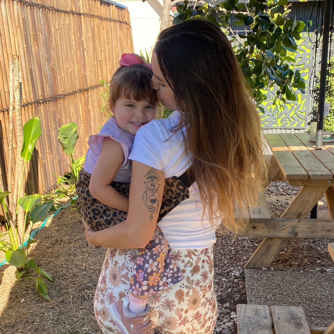 Woman holding a young child outdoors with plants and a wooden bench in the background