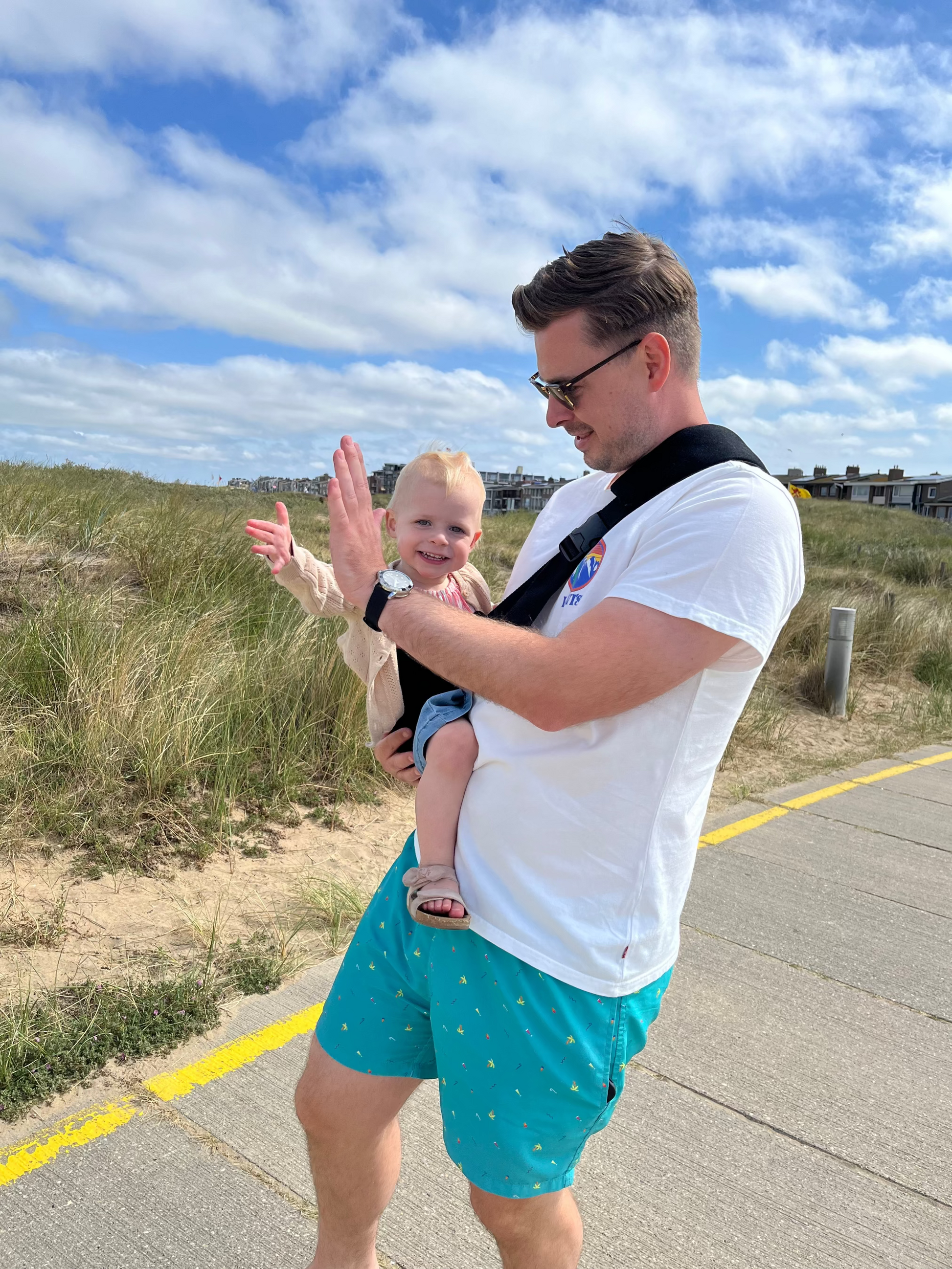 Man carrying a child on a beach path with grass and blue sky in the background